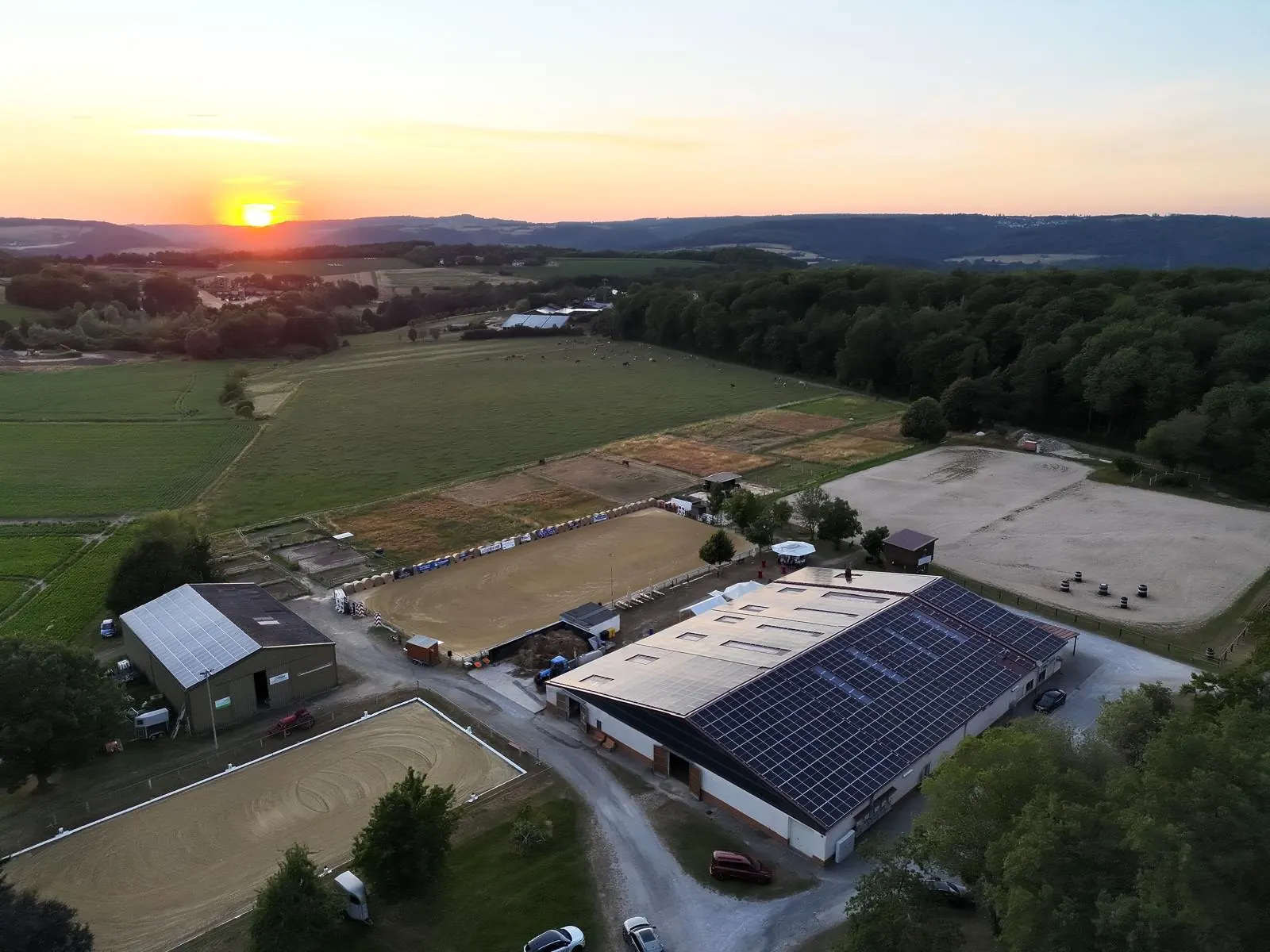 Luftbild der Reitanlage im Sonnenuntergang mit Reithalle, Solardach und mehreren Sandplätzen am Waldrand