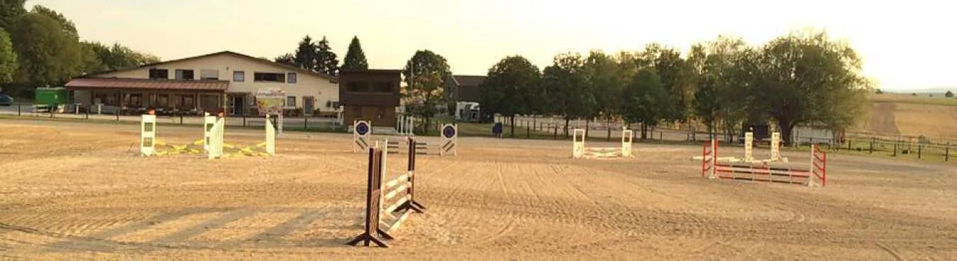 Sandplatz mit Hindernissen im Abendlicht, dahinter die Gebäude der Reitanlage